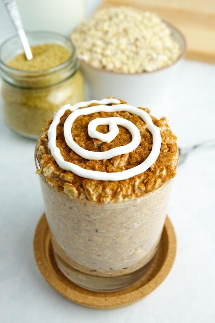 Photo of cinnamon roll overnight oats in a glass on a counter with ingredients like oats and flaxseed. 