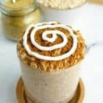 Cup filled with cinnamon roll overnight oats sitting on a coaster on a white marble counter, with ingredients displayed in the background.