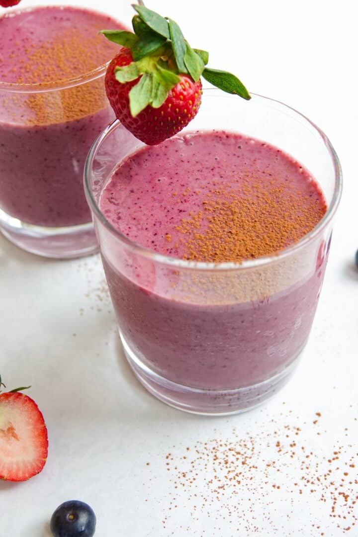 Photo of two chocolate berry smoothies in clear glasses sitting on a white marble counter and dusted with cocao powder.