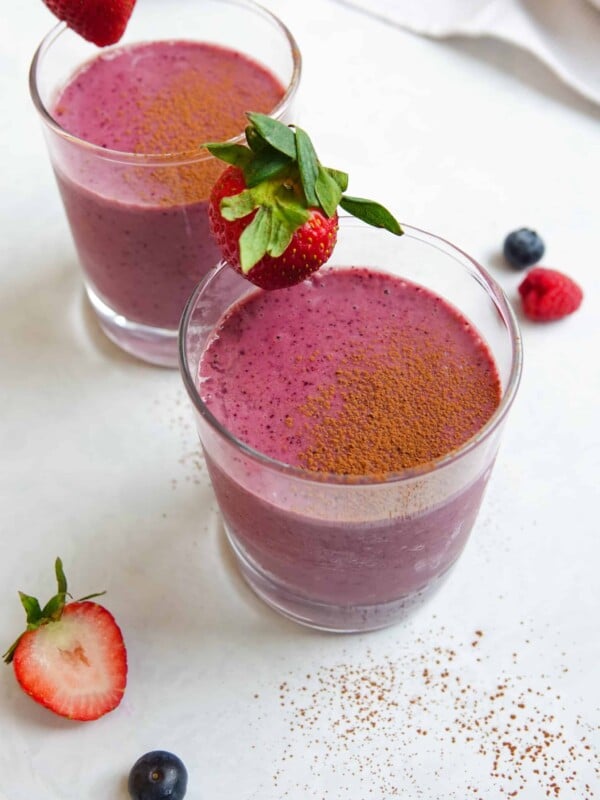 Photo of two chocolate berry smoothies in clear glasses sitting on a white marble counter and dusted with cocao powder.
