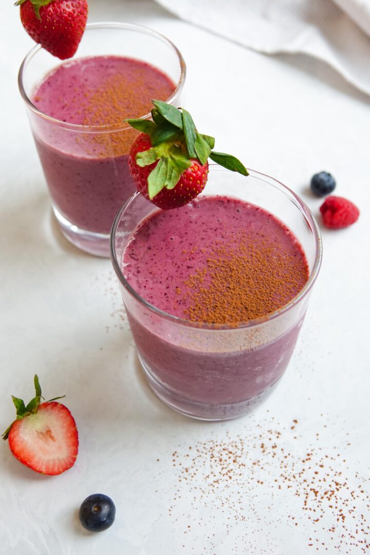 Photo of two chocolate berry smoothies in clear glasses sitting on a white marble counter and dusted with cocao powder.