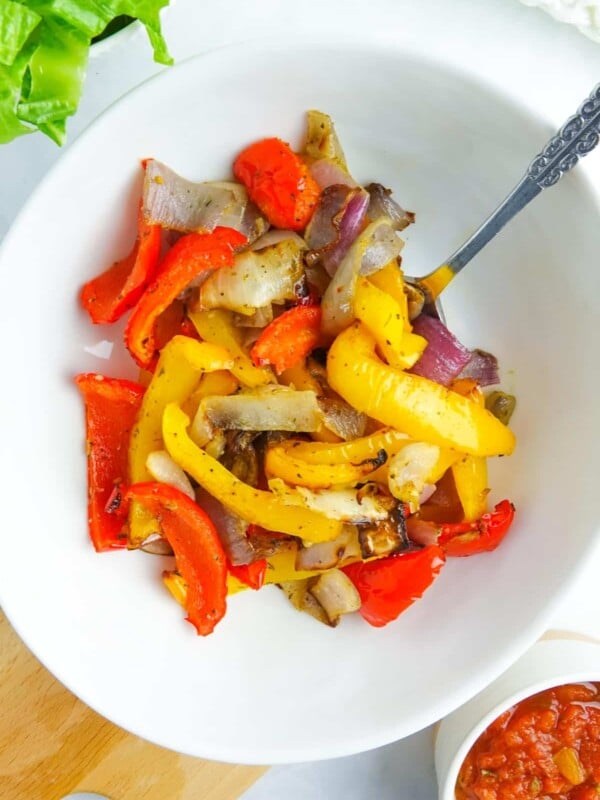 Photo of cooked peppers and onions in a white bowl, displayed over a wooden cutting board.