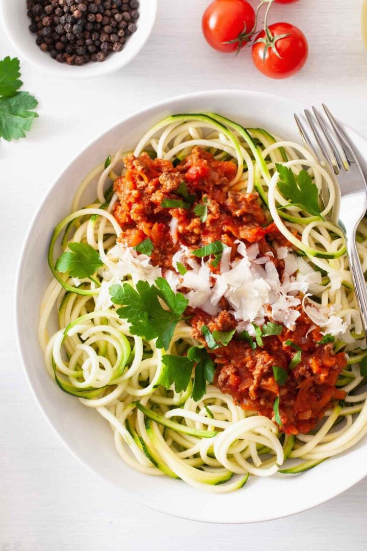 Bowl of zucchini noodles with meat sauce and grated parmesan cheese, displayed on a white counter with ingredients.