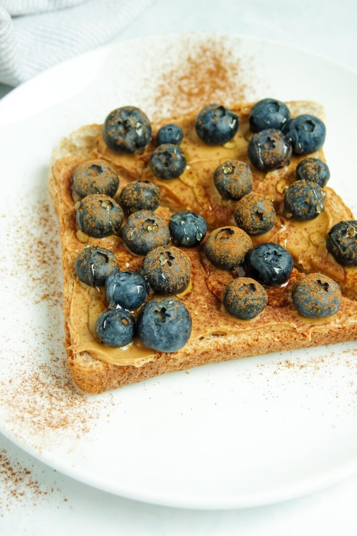 A close-up photos of slice of whole wheat toast cut in half, with peanut butter, blueberries, cinnamon and honey displayed on a white plate on a white marble counter.