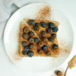 A slice of whole wheat toast with peanut butter, blueberries, cinnamon and honey displayed on a white plate on a white marble counter.