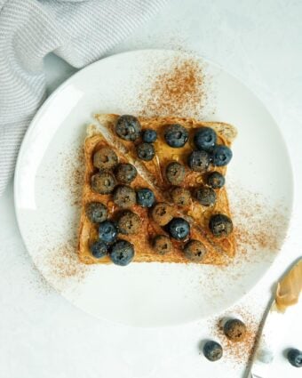 A slice of whole wheat toast with peanut butter, blueberries, cinnamon and honey displayed on a white plate on a white marble counter.