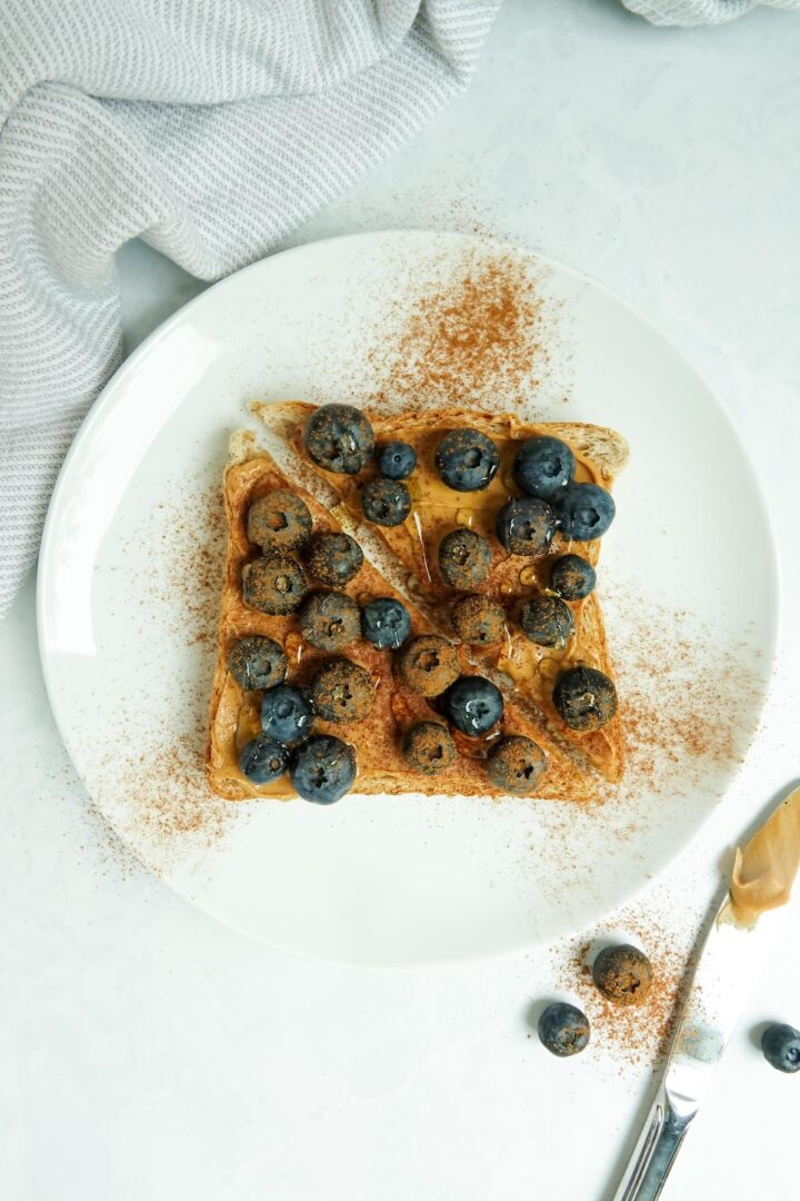 A slice of whole wheat toast with peanut butter, blueberries, cinnamon and honey displayed on a white plate on a white marble counter.