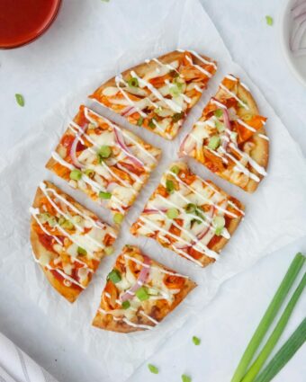 Whole buffalo chicken flatbread topped with green onions, red onions, and a drizzle of sour cream displayed on parchment paper on a white counter