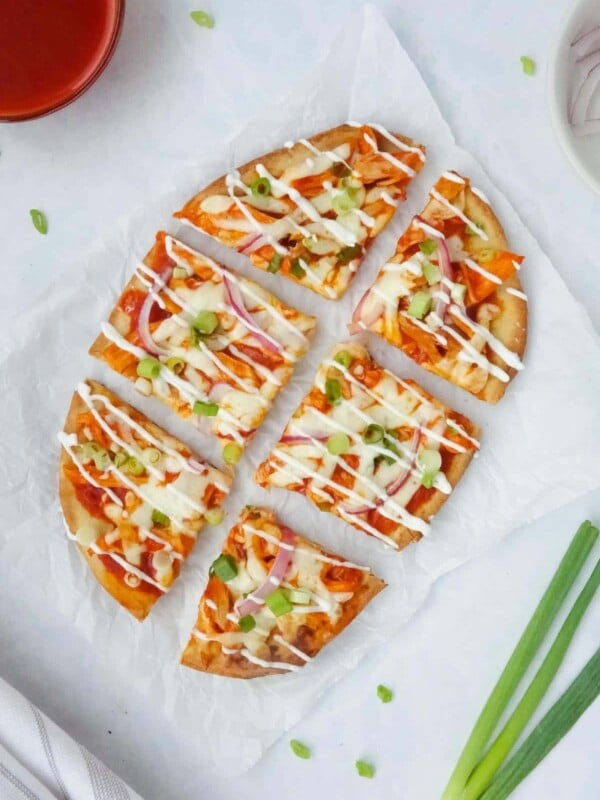 Whole buffalo chicken flatbread topped with green onions, red onions, and a drizzle of sour cream displayed on parchment paper on a white counter