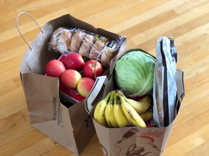 Paper grocery bags sitting on a hardwood floor filled with healthy foods like bananas, apples and bread.