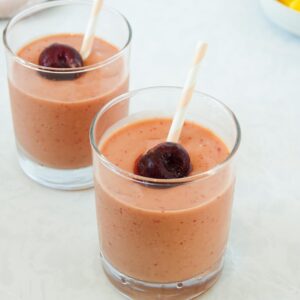 Photo of two mango cherry smoothies in small clear glasses with pink and white striped straws, displayed on a white marble counter.