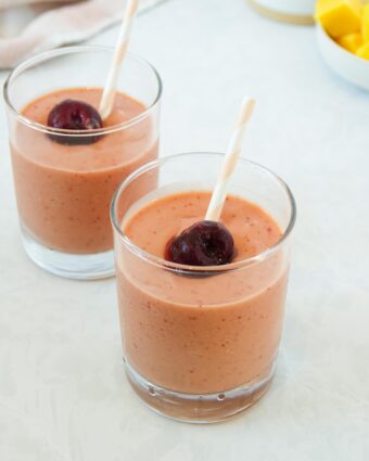 Photo of two mango cherry smoothies in small clear glasses with pink and white striped straws, displayed on a white marble counter.
