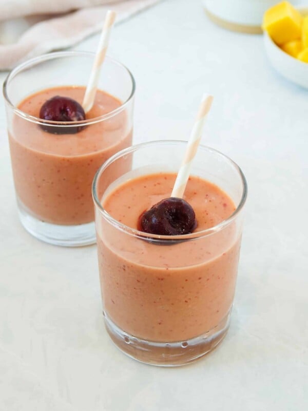 Photo of two mango cherry smoothies in small clear glasses with pink and white striped straws, displayed on a white marble counter.