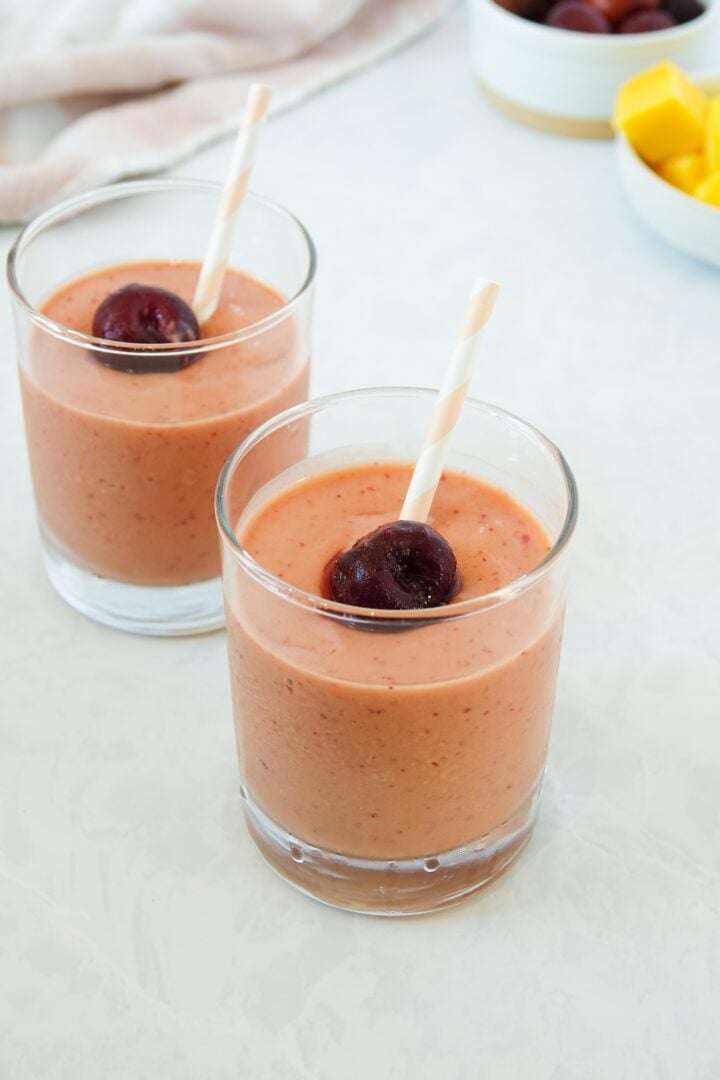 Photo of two mango cherry smoothies in small clear glasses with pink and white striped straws, displayed on a white marble counter. 