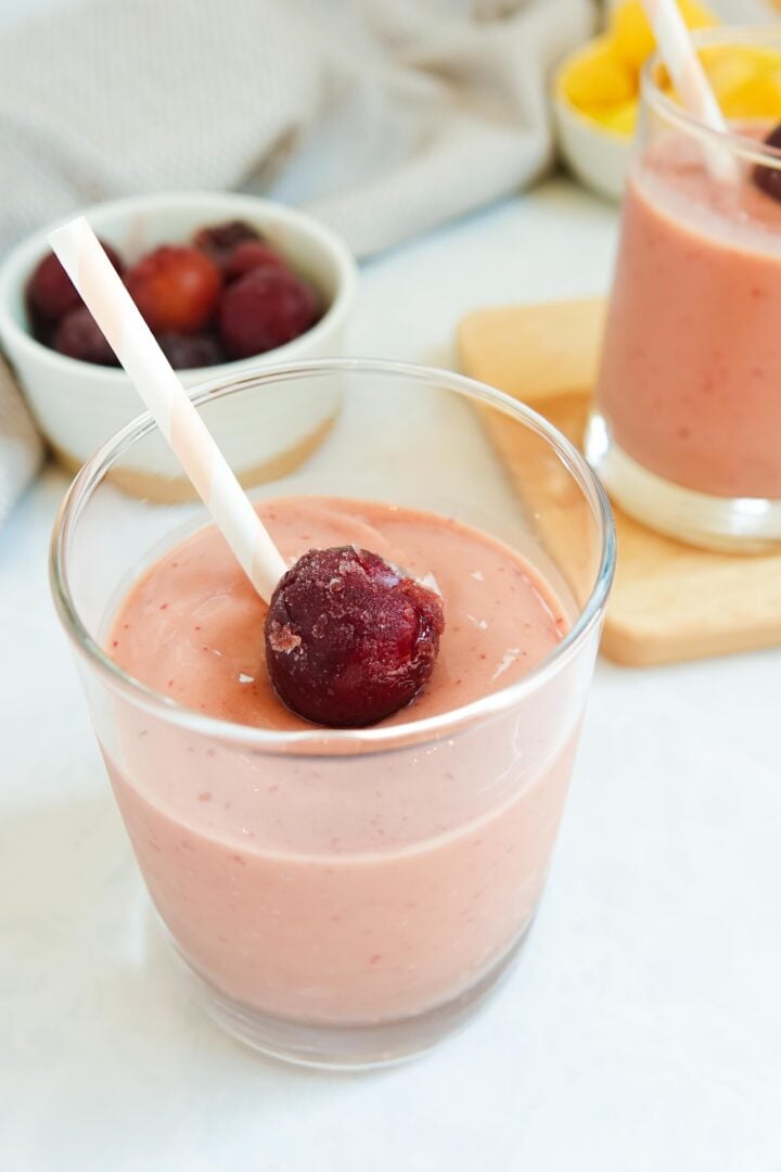 Mango cherry smoothie in a small glass topped with a frozen cherry and a pink and white striped straw.