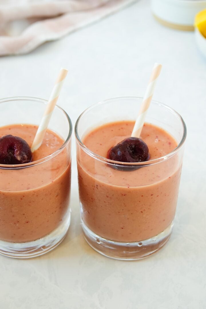 Two glasses of mango cherry smoothie topped with a frozen cherry and a pink and white striped straw, displayed side-by-side on a white marble counter. 