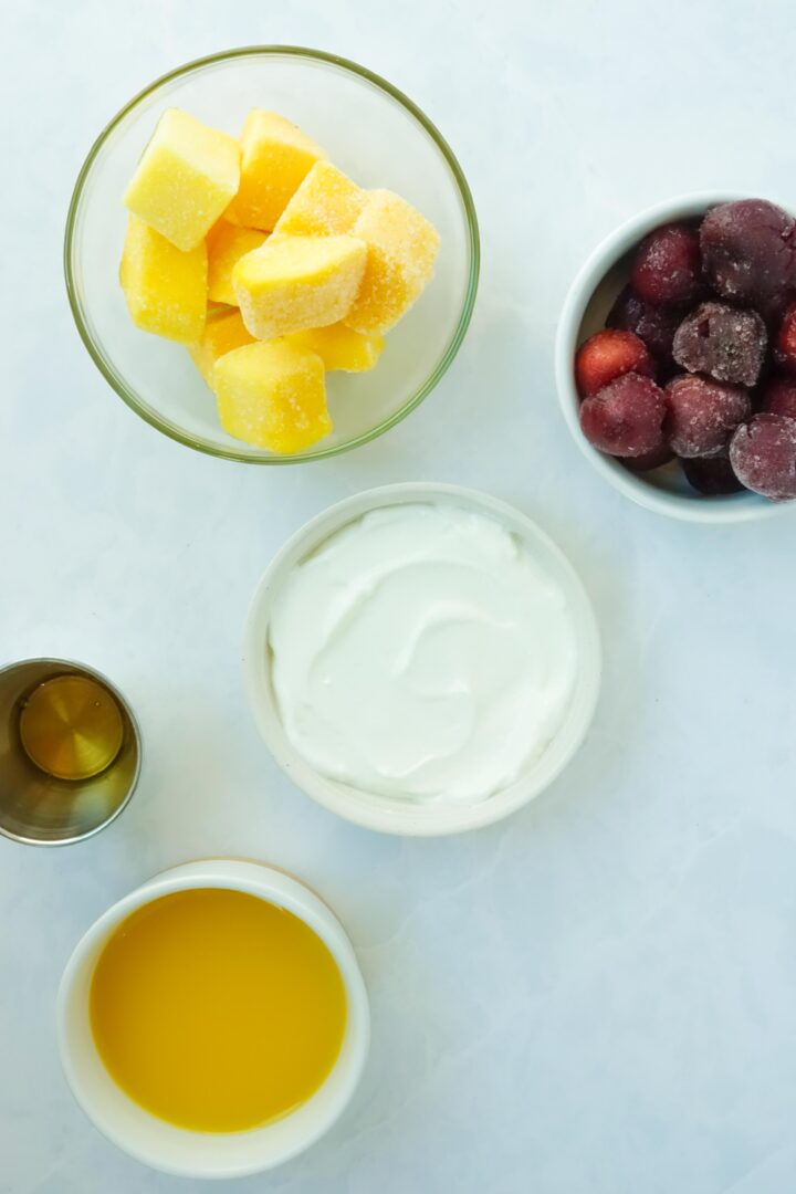 Ingredients for mango cherry smoothie displayed in small bowls on a white marble counter, including frozen pineapple, frozen mango, greek yogurt, orange juice, and honey.