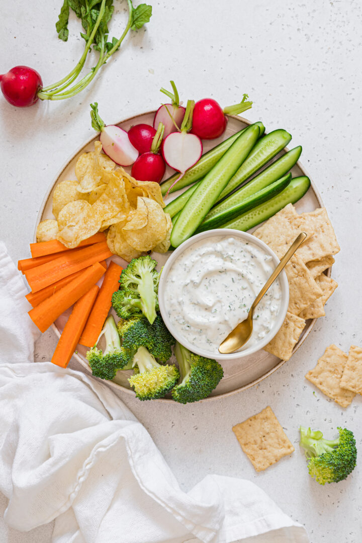 Healthy ranch dip with greek yogurt in a small white bowl, plated with assorted raw vegetables, crackers, and potato chips.