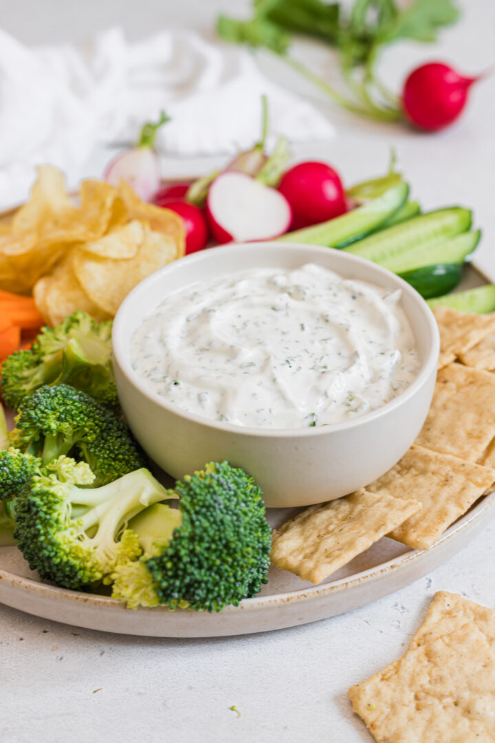 Bowl of healthy ranch dip with greek yogurt displayed in a small white bowl, served with assorted raw vegetables, crackers, and chips.