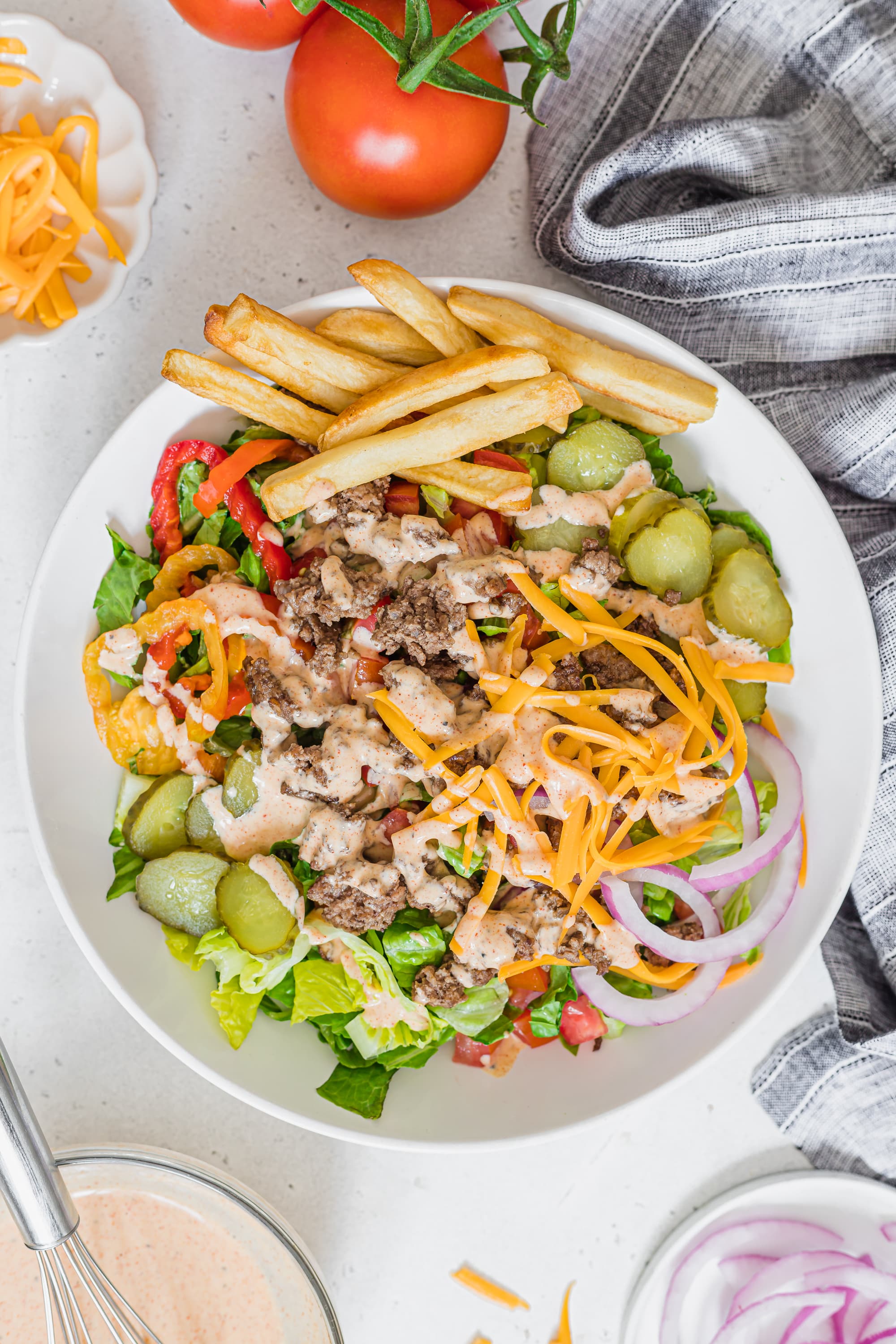 Assembled and un-mixed burger salad on a white kitchen counter surrounded by ingredients and a grey tea towel.