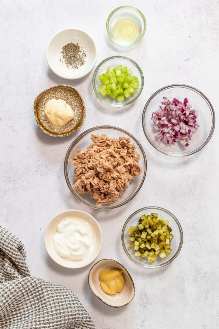 Ingredients for dill pickle tuna salad displayed in small bowls on a white countertop.