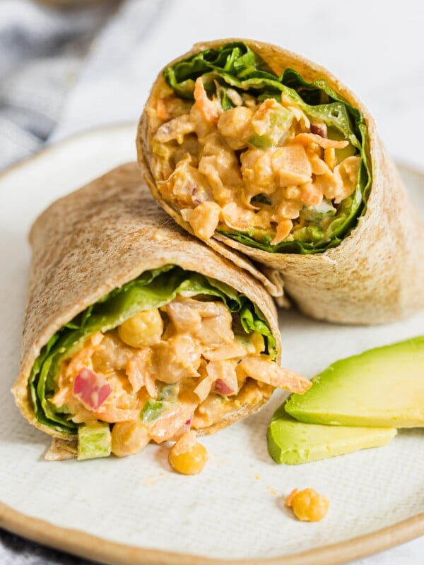 Photo of buffalo chickpea salad wraps cut in half and displayed on a white plate with a few slices of avocado.