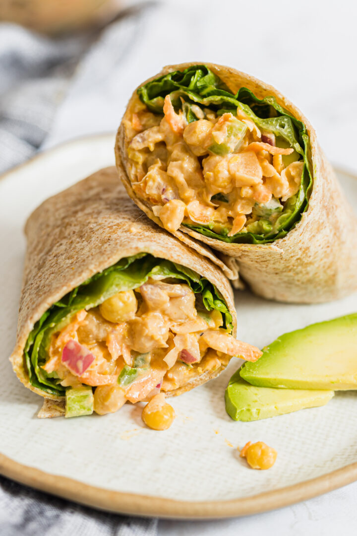 Photo of buffalo chickpea salad wraps cut in half and displayed on a white plate with a few slices of avocado.