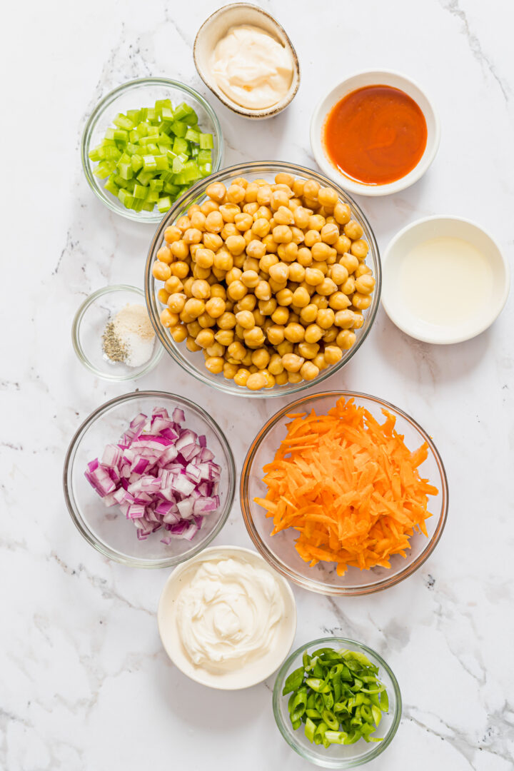 Ingredients to make buffalo chickpea salad wraps displayed in bowls on a white marble counter.