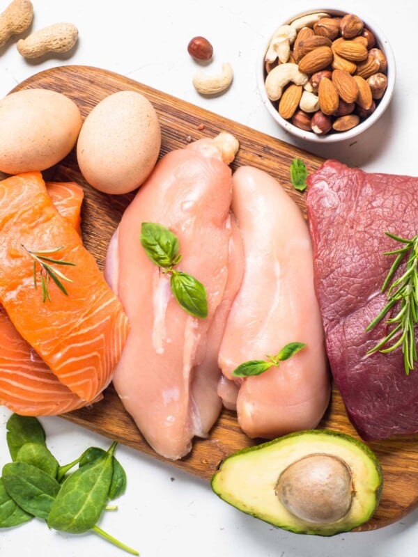 Flatlay photo of high protein foods displayed on a wood board on a white counter, including raw chicken, raw salmon and eggs.