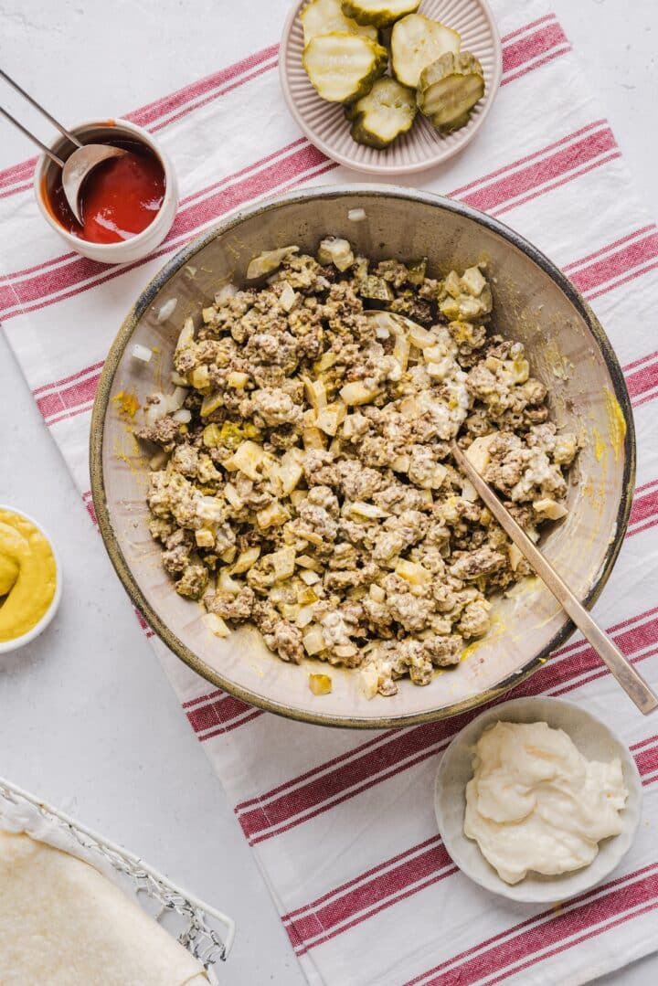 Ground beef in a large mixing bowl with condiments, diced onion, and diced pickle.