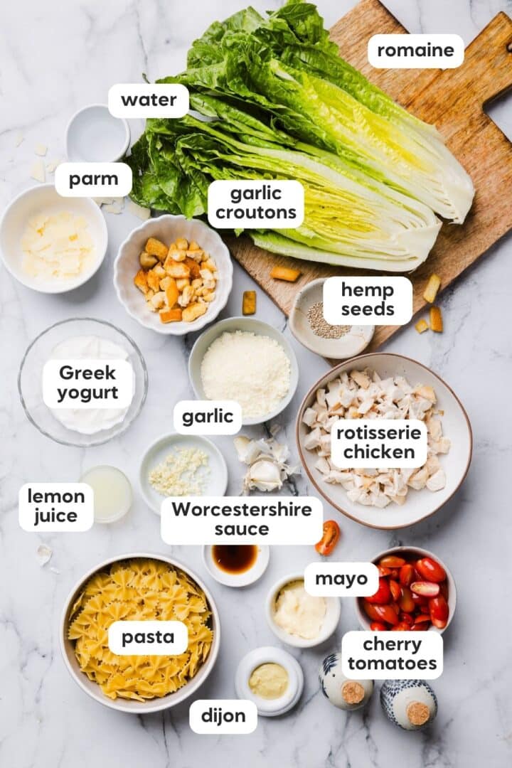 Ingredients for chicken caesar pasta salad displayed in small bowls on a grey marble counter.