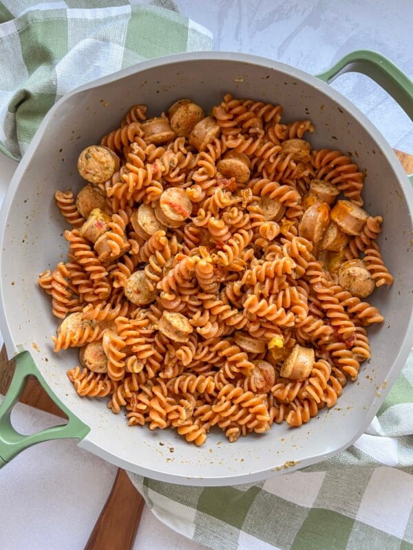 Large green pan with pasta and sausage displayed on a wood board with a green checkered kitchen cloth