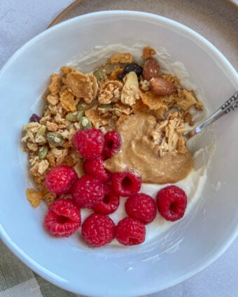 high protein yogurt bowl in a white bowl, topped with raspberries, peanut butter, and granola