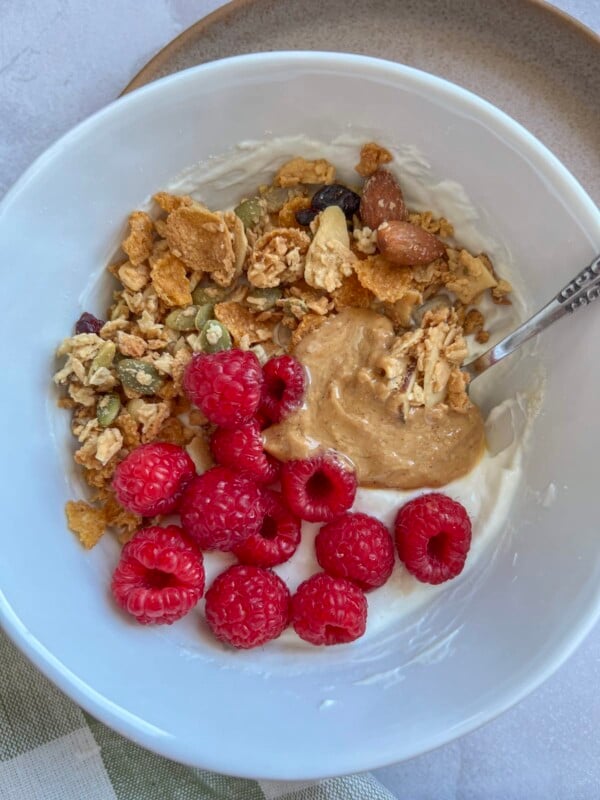 high protein yogurt bowl in a white bowl, topped with raspberries, peanut butter, and granola