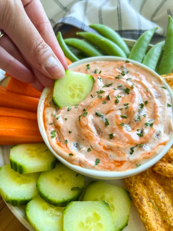 Woman's hand dipping a cucumber in a small bowl of salsa ranch dressing, surrounded by veggies and chips