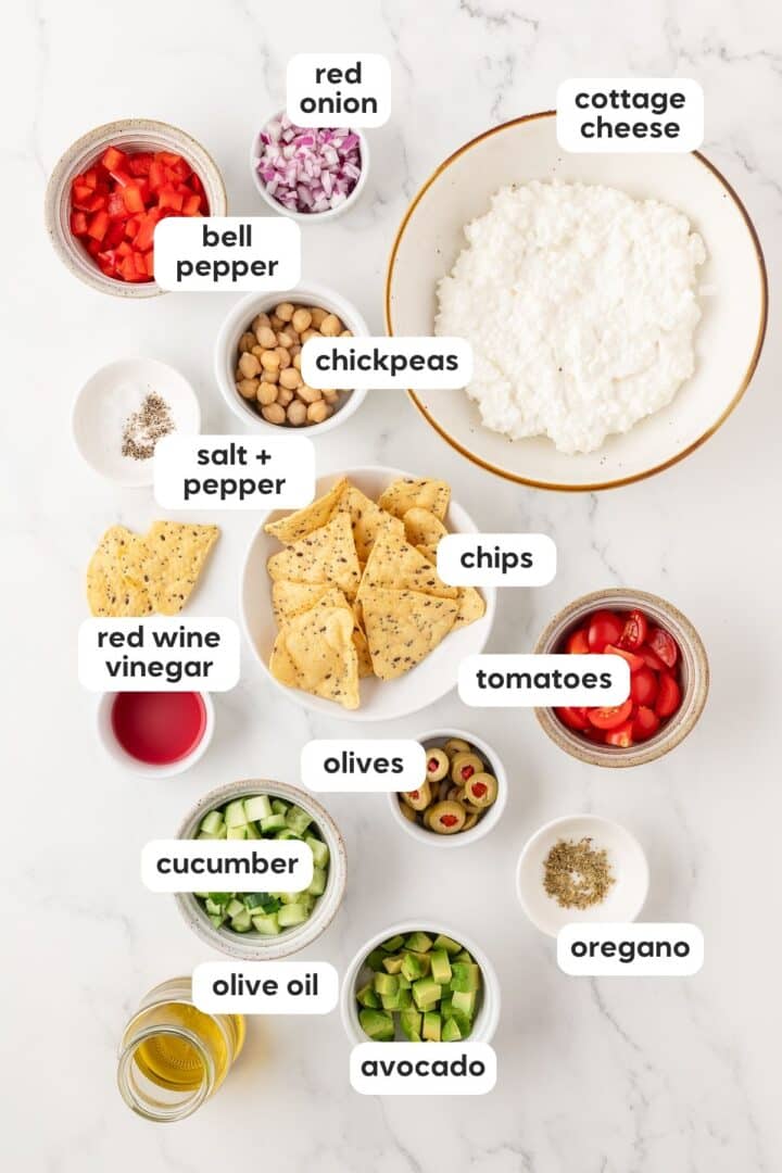 Ingredients for cottage cheese greek salad displayed in small bowls on a white marble counter.