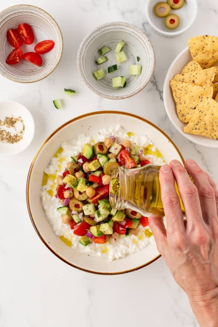 Woman's hand pouring olive oil over a cottage cheese greek salad in a white bowl.