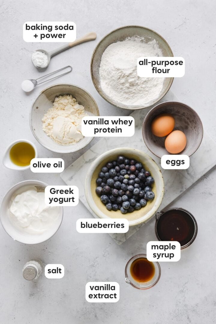 Ingredients for high protein blueberry muffins displayed in small bowls on a grey marble counter top.