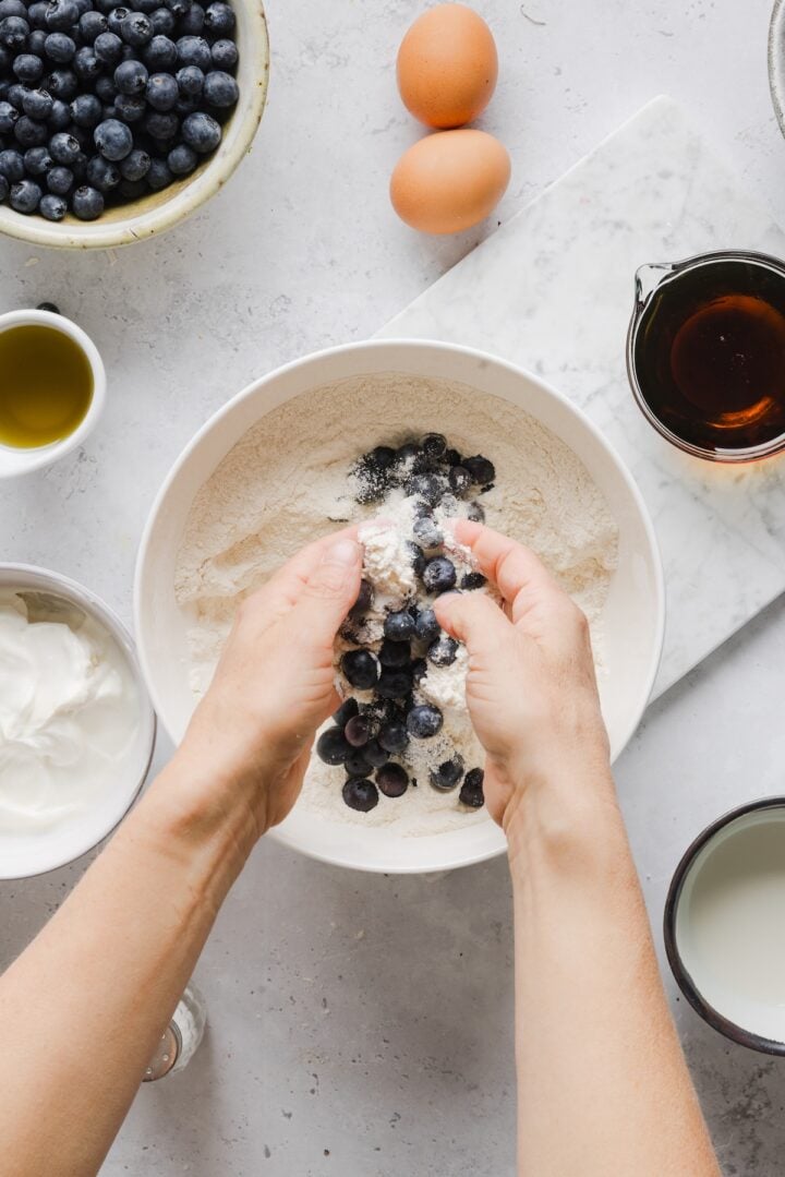 Flour and fresh blueberries being tossed together by a woman's hand in a large white bowl.