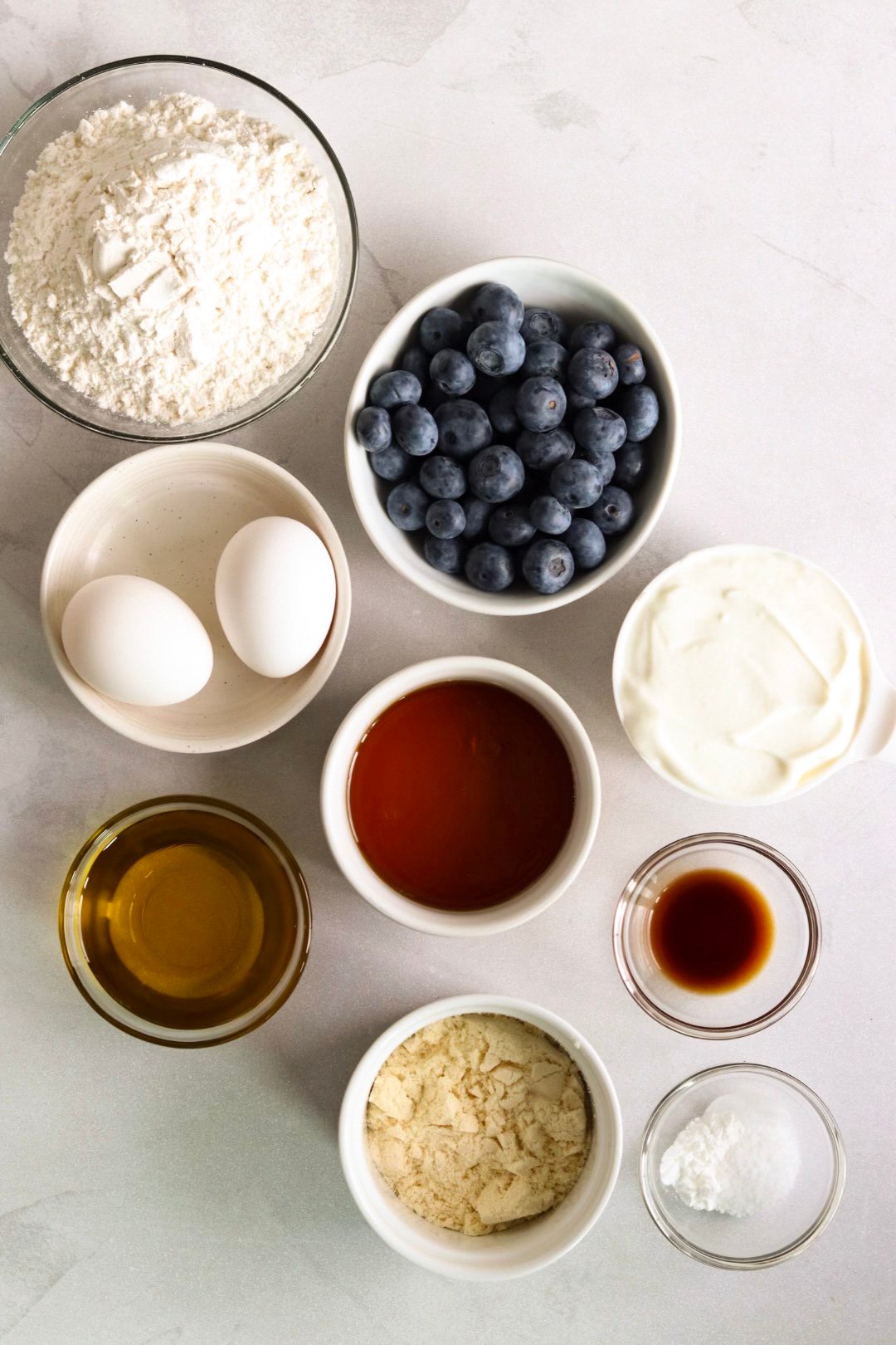 Ingredients for high protein blueberry muffins displayed in bowls on a white kitchen counter