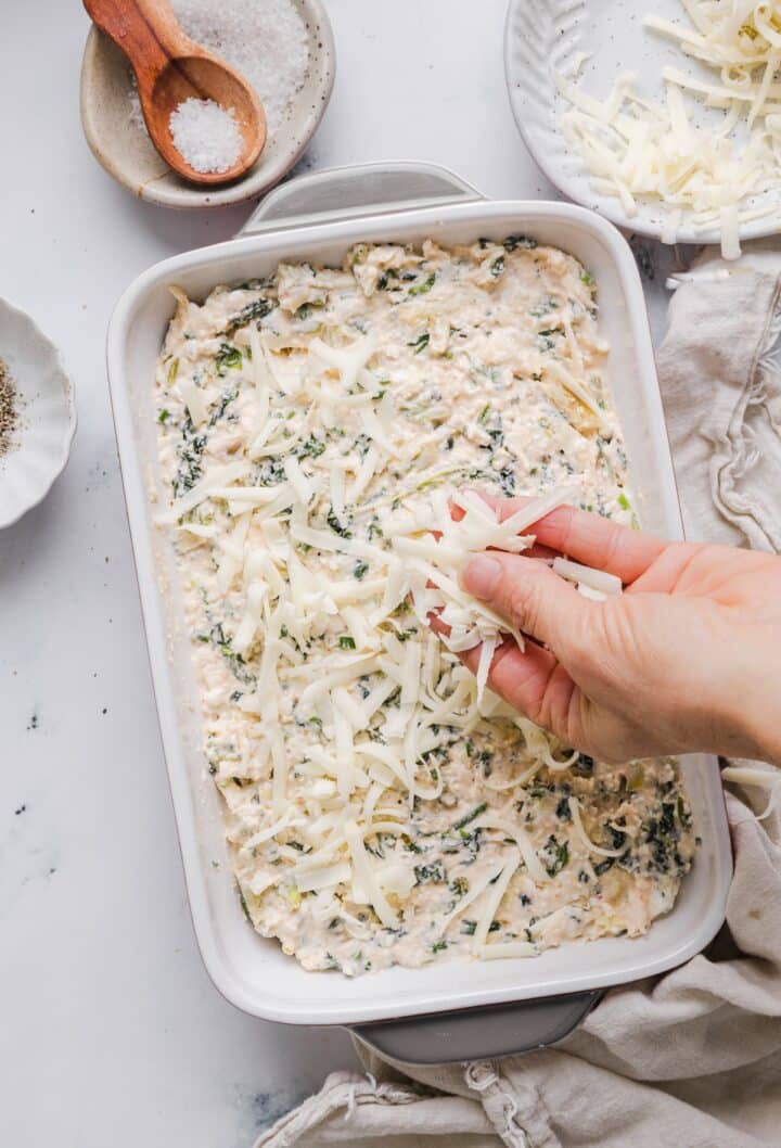 Healthy spinach artichoke dip in a rectangular baking dish, being topped with cheese.