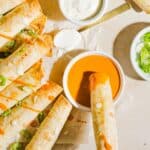 Baked buffalo chicken taquitos lined up on parchment paper, with a woman's hand dipping into buffalo sauce