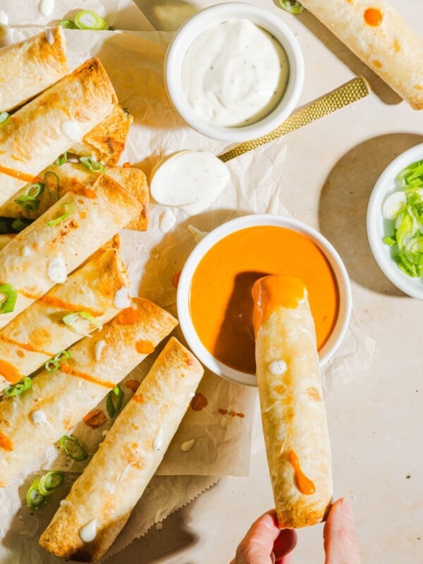 Baked buffalo chicken taquitos lined up on parchment paper, with a woman's hand dipping into buffalo sauce