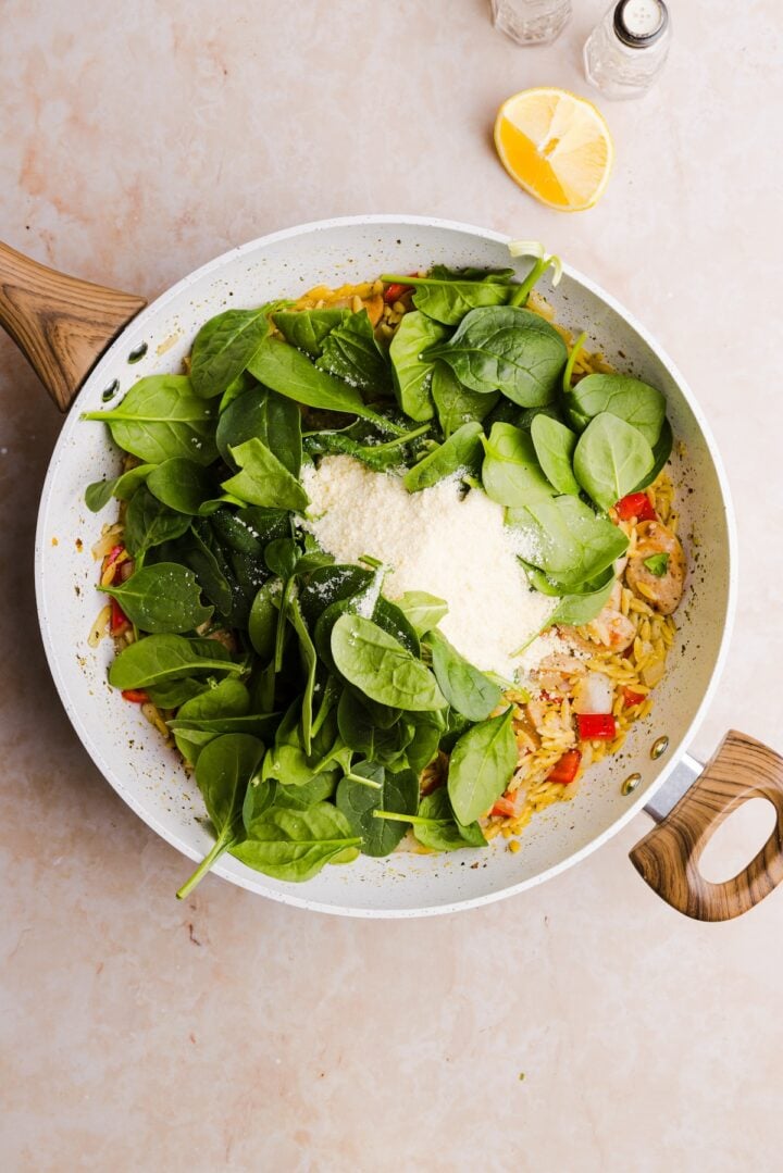 Spinach and parmesan added to a large white pan with other ingredients for an orzo dish