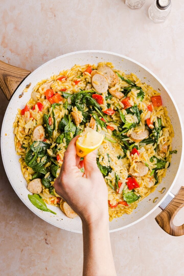 A women's hand squeezing fresh lemon juice over a cooked skillet with chicken sausage, orzo and other veggies.