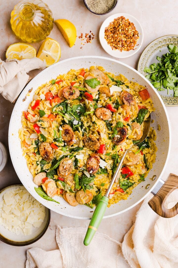Overhead photo of chicken sausage orzo styles in a large white pan, with a silver serving spoon with a green handle