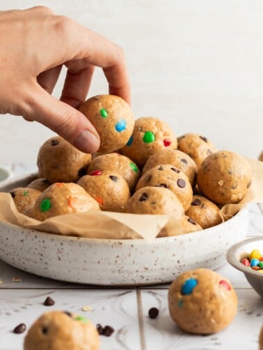 Cookie dough protein balls in a large white bowl, with a woman's hand grabbing one to eat