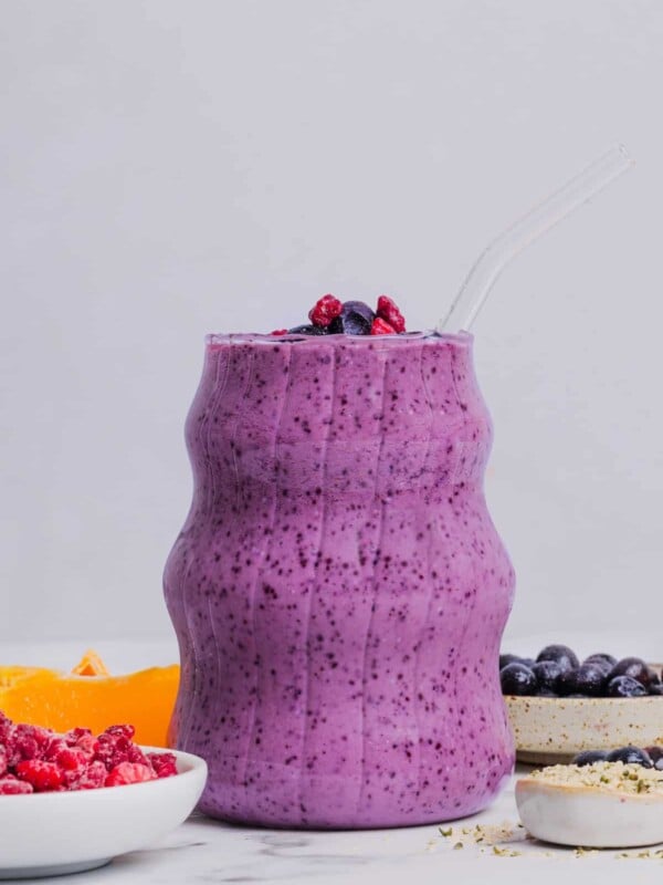 Blueberry raspberry smoothie in an etched glass sitting near fruit and other ingredients