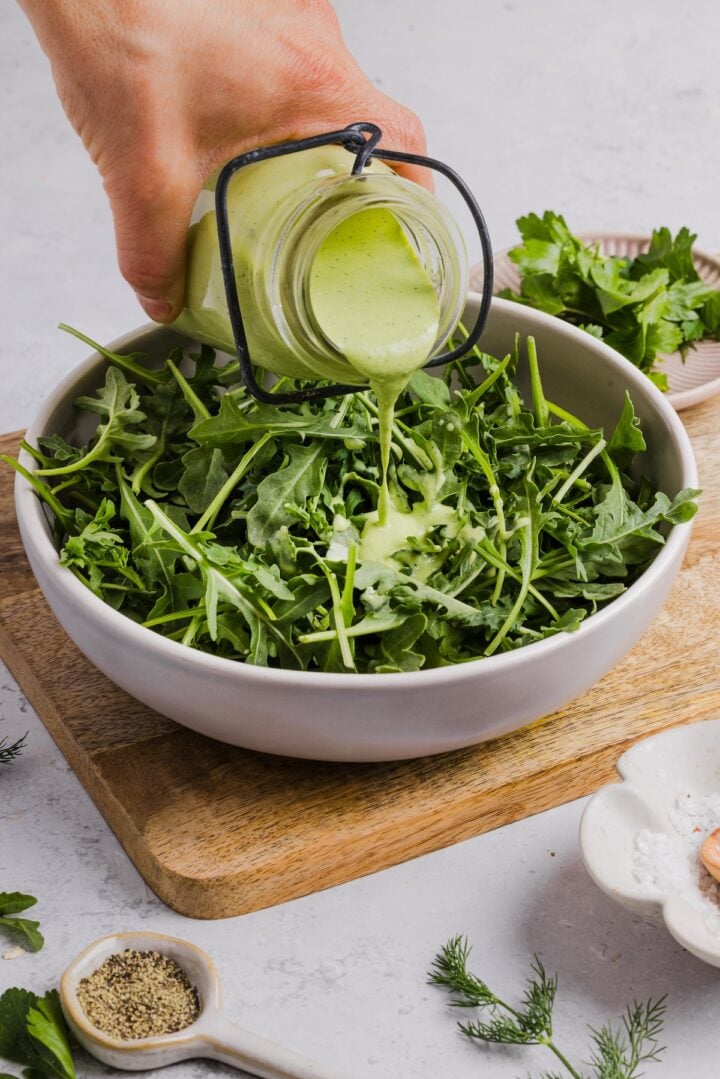 Green goddess ranch salad dressing being poured over arugula and greens in a shallow white bowl