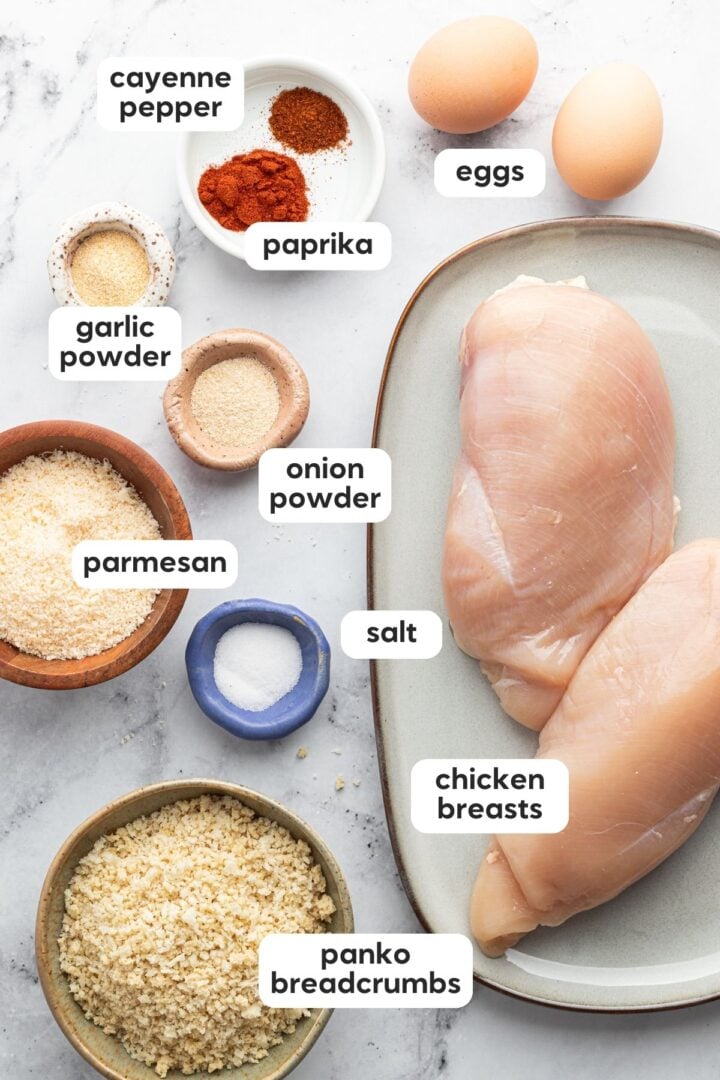 Labeled photo of ingredients for crispy, baked, panko chicken nuggets, displayed in small bowls on a marble kitchen counter